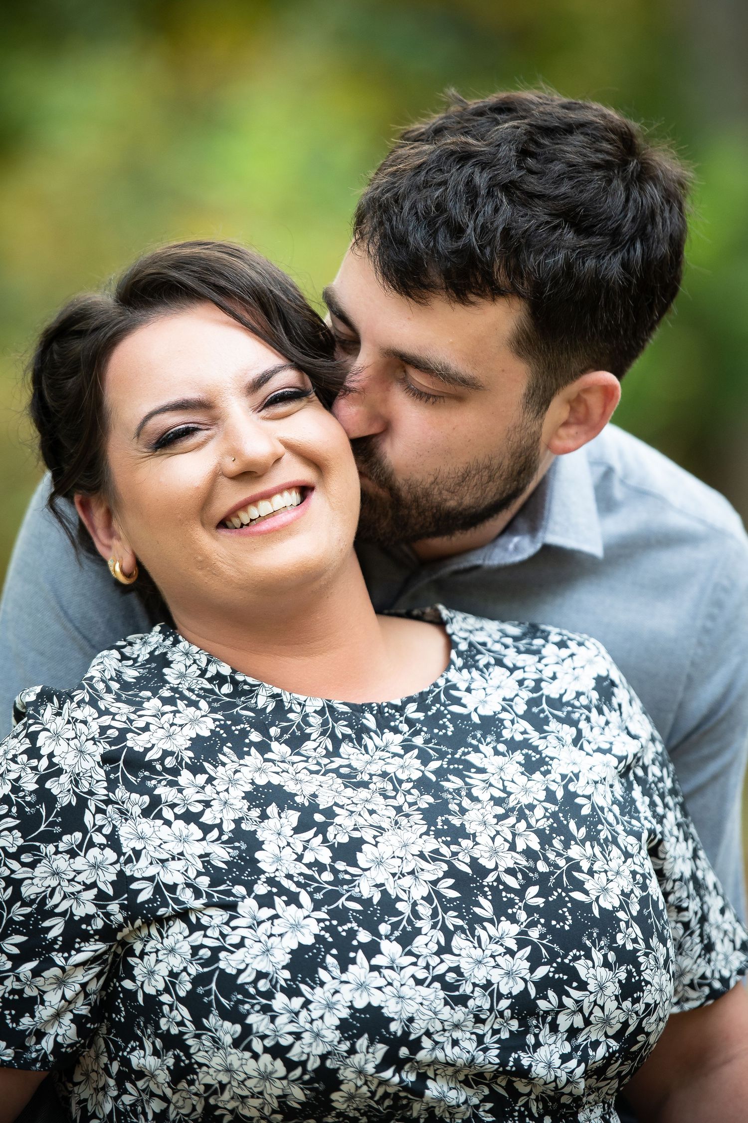 A smiling woman in a floral dress receives a tender kiss on the cheek from a bearded man outdoors.