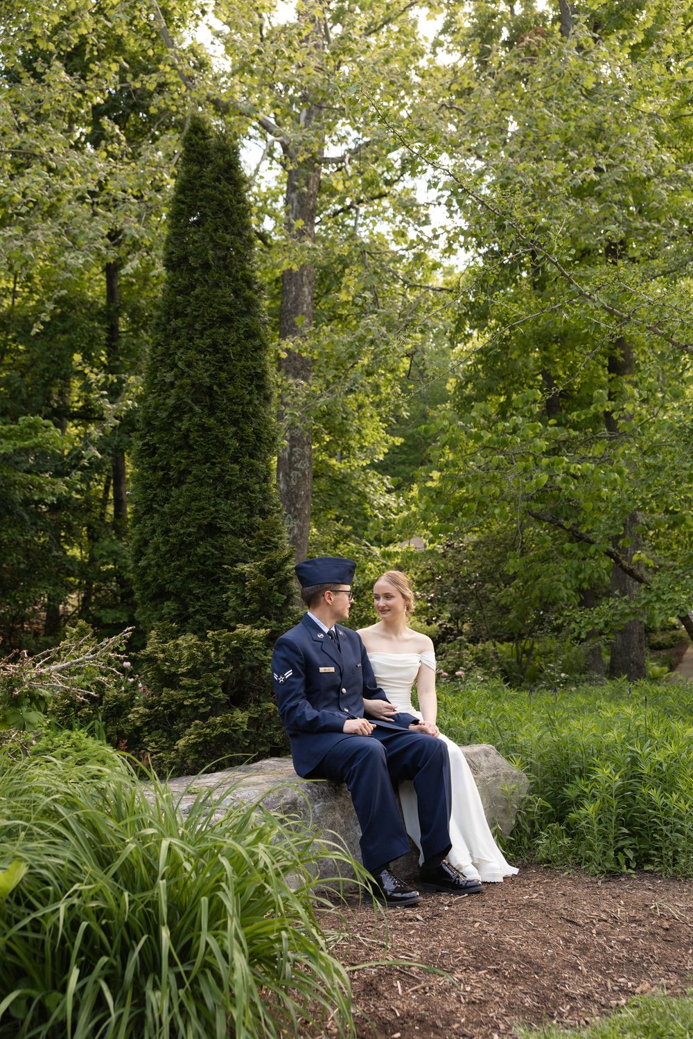 Military groom and bride in white dress sitting on a rock surrounded by lush green trees in an outdoor garden setting.