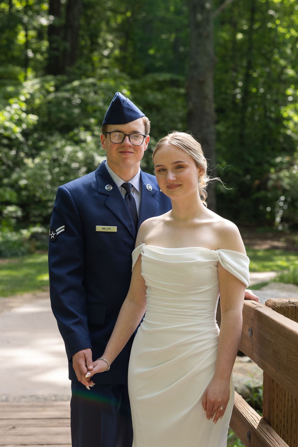Military groom in Air Force dress uniform poses with bride in white off-shoulder wedding gown on wooden bridge.