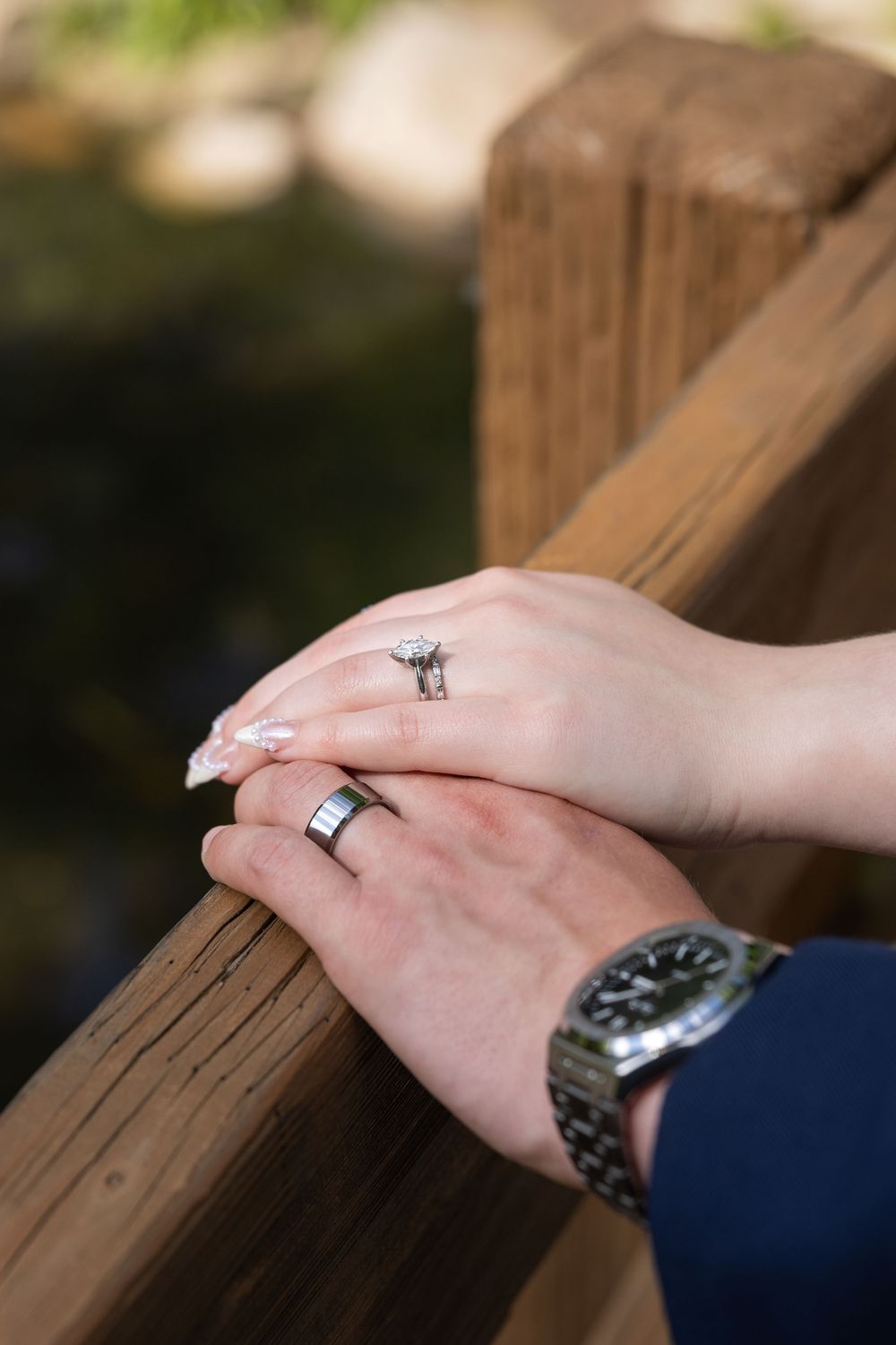 Couple's hands resting on wooden railing, wearing wedding rings and a wristwatch, symbolizing love and commitment.