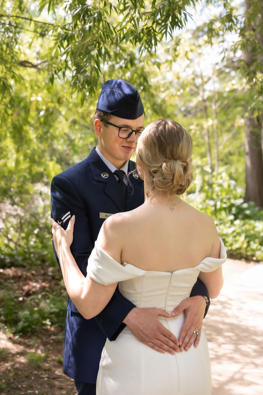 Military groom in blue uniform embraces bride in off-shoulder wedding dress outdoors among lush green trees.