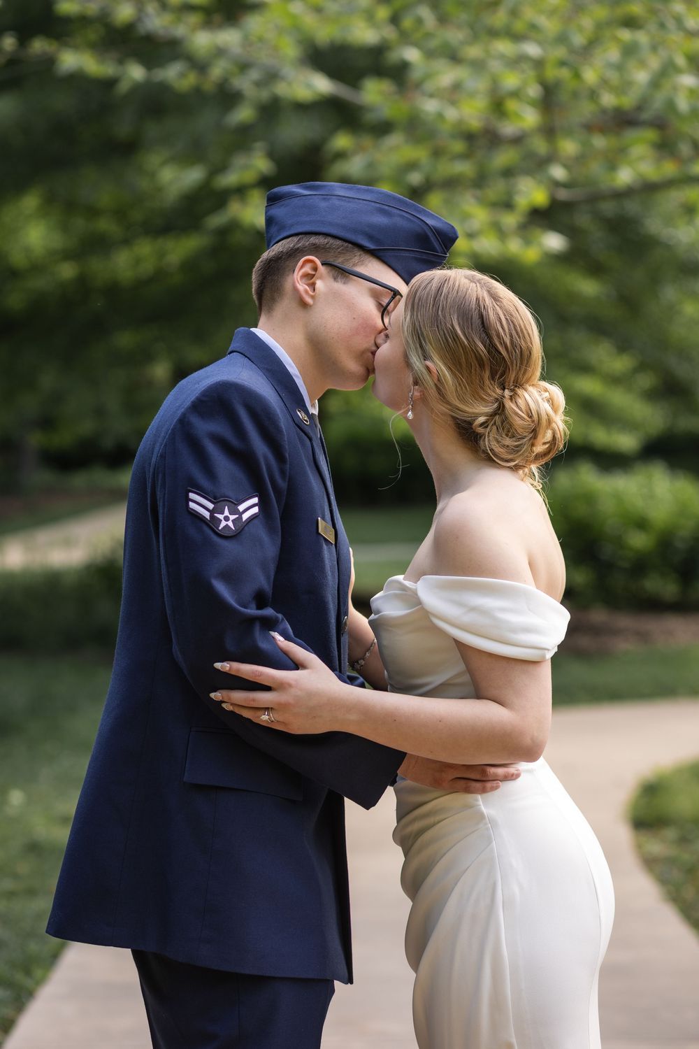 Military groom in Air Force uniform kissing bride in white off-shoulder gown outdoors among lush green trees.