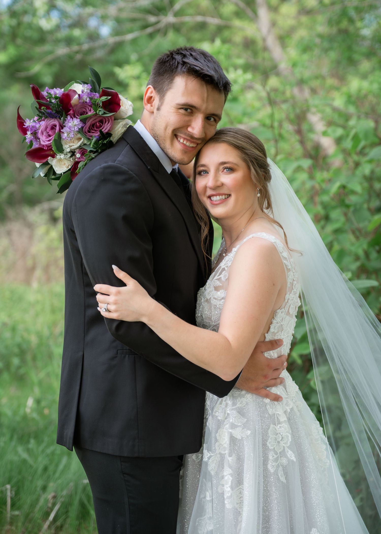 A smiling bride and groom embrace outdoors, she in a lace gown with veil, he in a black suit, holding a red bouquet.