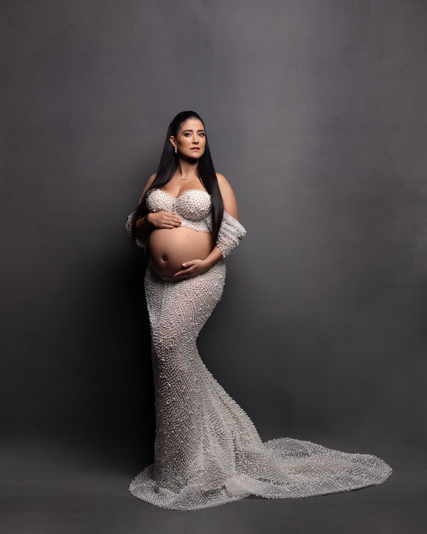 A maternity photoshoot featuring a flowing silver beaded gown against a dark background.