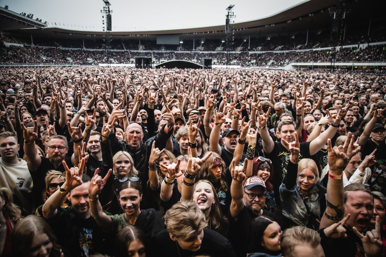 Massive crowd of rock fans throwing devil horns at an outdoor stadium concert.