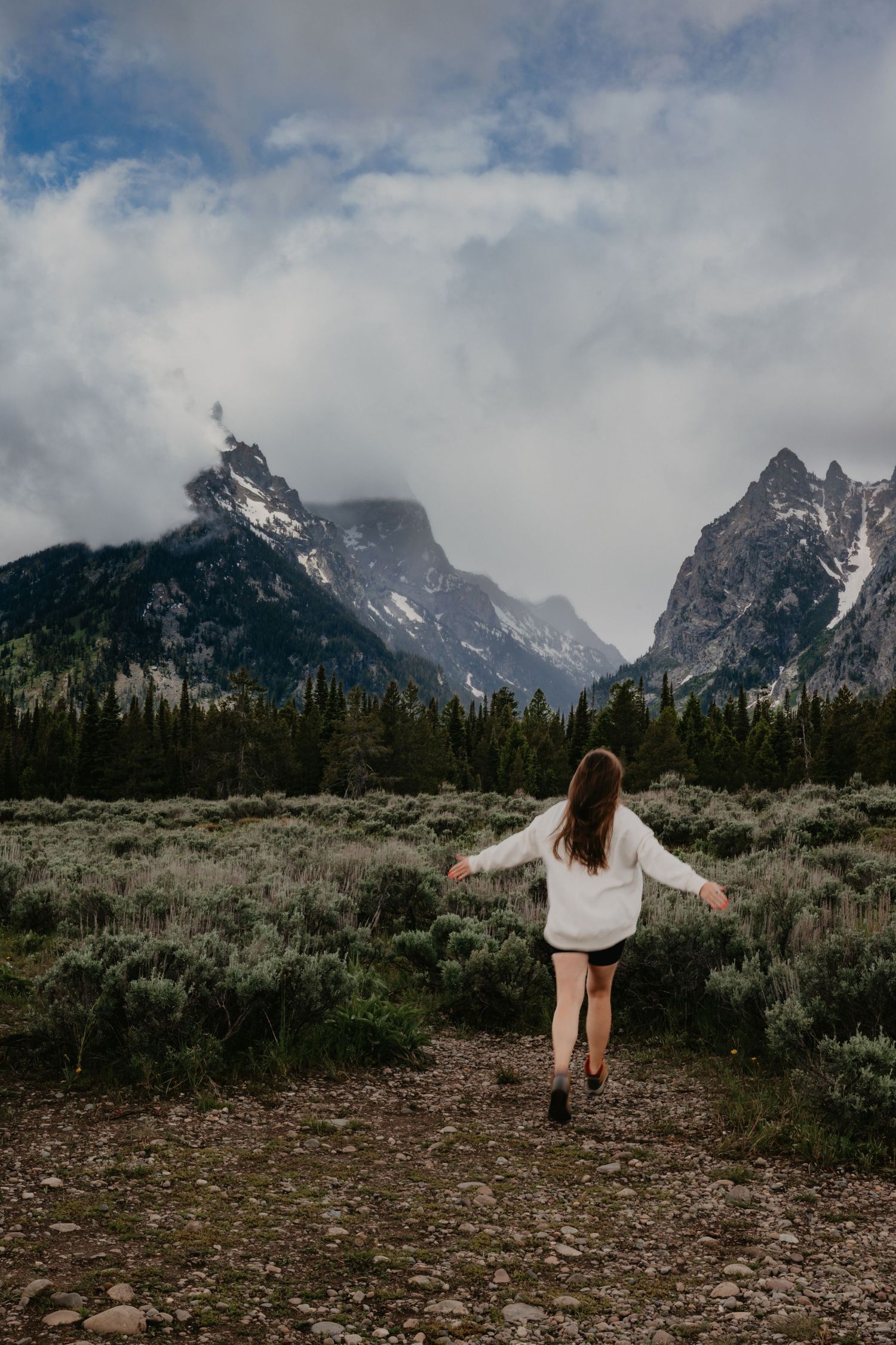Woman in white sweater walks toward dramatic snow-capped Grand Teton mountains under cloudy sky.
