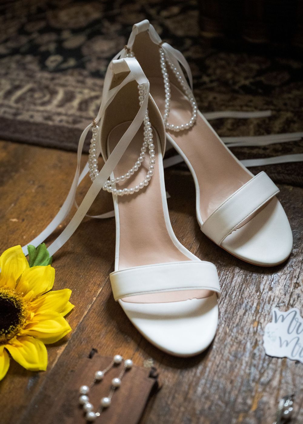 Elegant white bridal heels with pearl ankle straps displayed beside a sunflower on rustic wood.