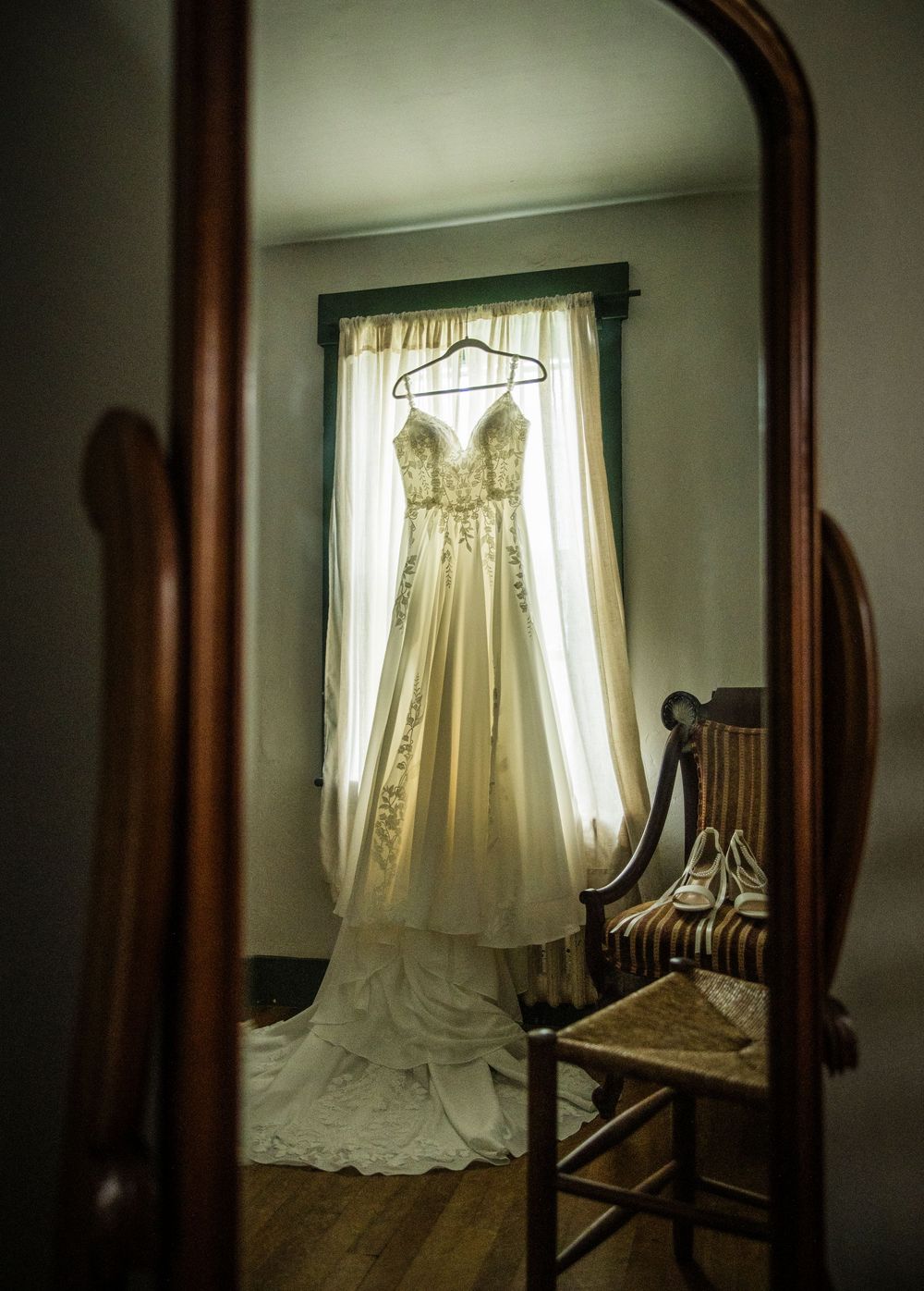 Elegant lace wedding dress hanging by a window reflected in a vintage wooden mirror in a softly lit room.