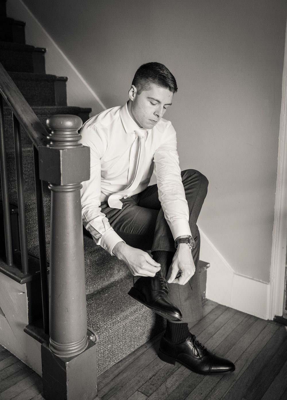 Young man in white dress shirt tying dress shoes on staircase in black and white wedding photo.