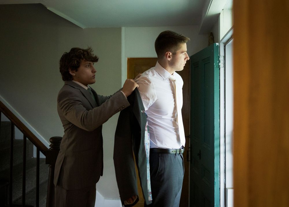 Groomsman helping groom put on suit jacket before wedding ceremony near a staircase.
