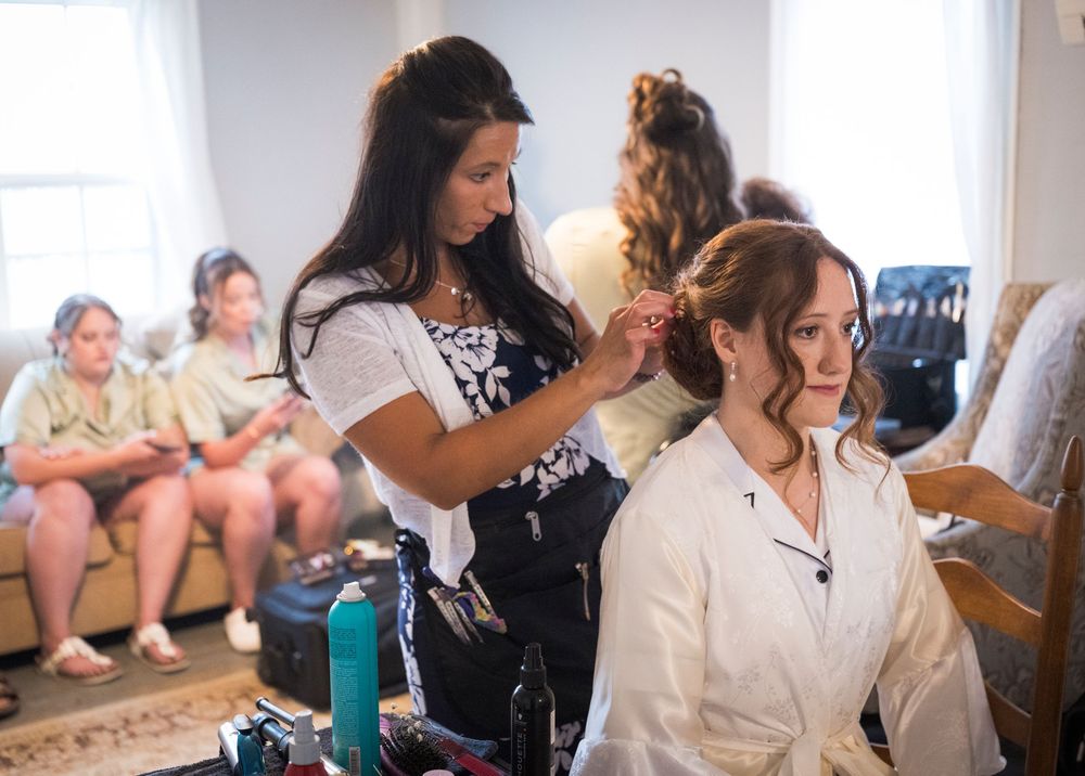 Hair stylist styling bride's hair while bridesmaids relax in background during wedding preparations.
