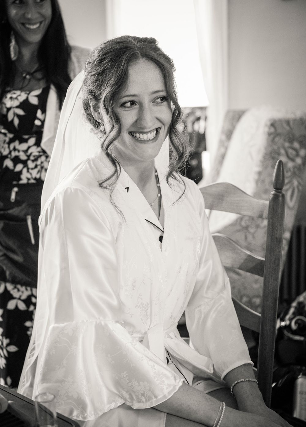 Radiant bride smiles in black and white photo wearing veil and white robe while getting ready.