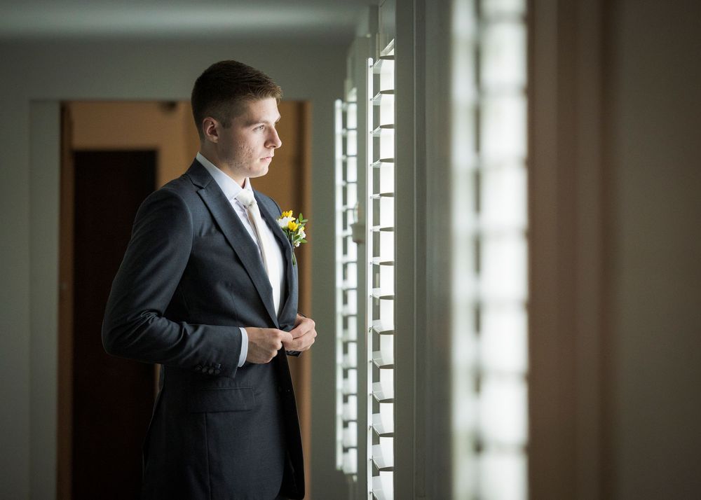 Dapper groom in dark suit with boutonniere gazing out window before wedding ceremony.