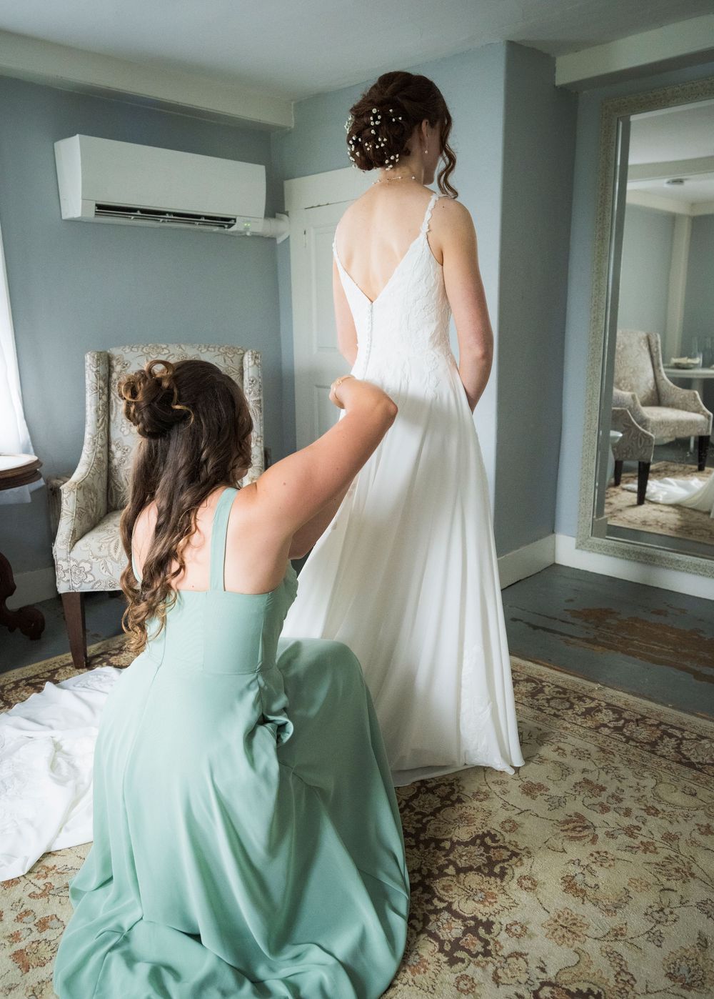 Bridesmaid in sage green dress kneeling to adjust bride's white wedding gown train in an elegant hotel room.