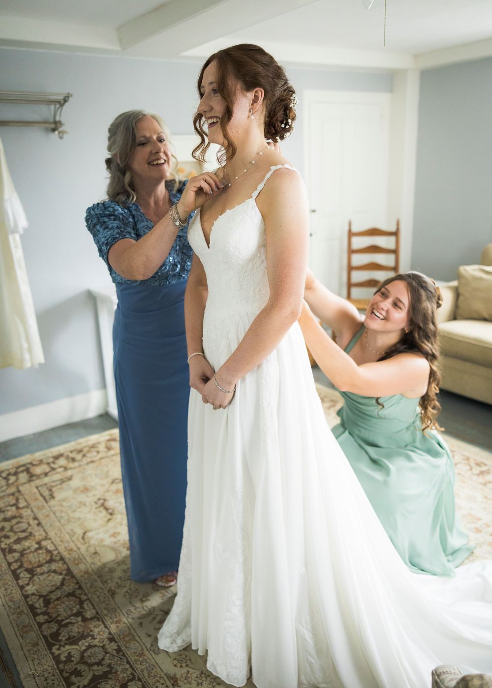 Bride being helped into white lace wedding gown by mother and smiling bridesmaid.