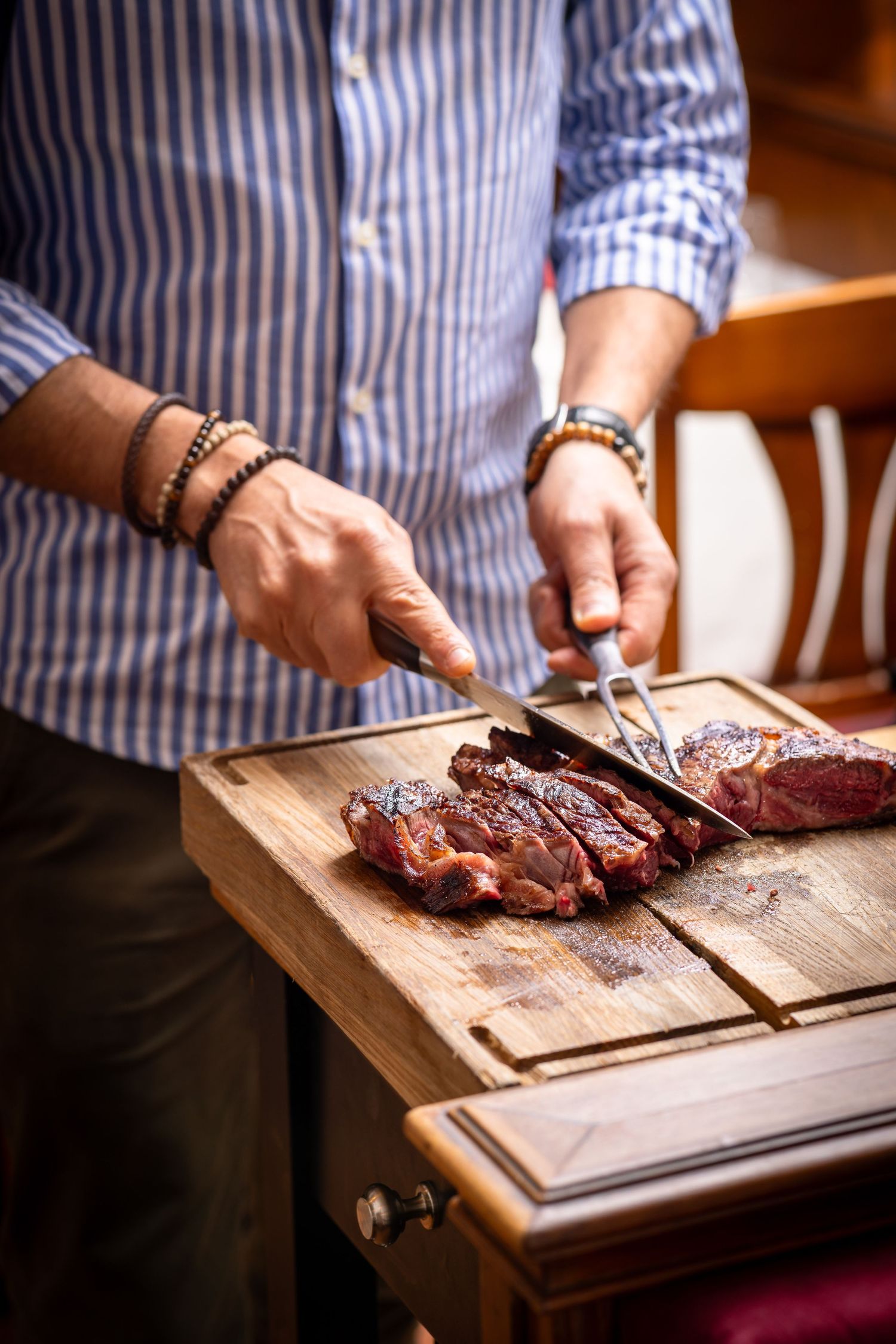 Man in striped shirt slicing grilled steak on wooden cutting board with knife and fork.
