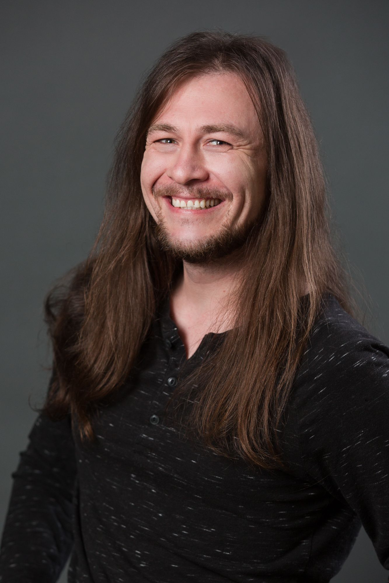 Smiling man with long brown hair and beard wearing a dark shirt, photographed against a gray studio background.