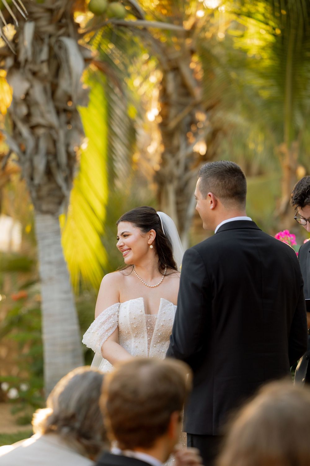 Smiling bride in a strapless lace gown during outdoor wedding ceremony among tropical palms.
