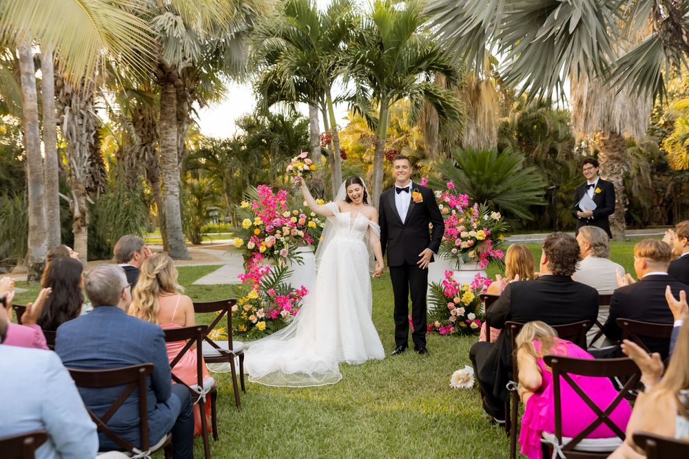 Bride and groom stand together at outdoor wedding ceremony surrounded by tropical palms and vibrant pink and yellow floral arrangements.