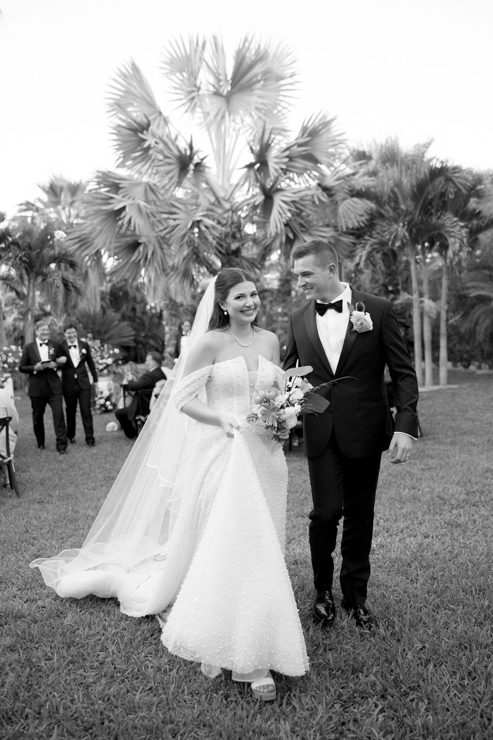 Black and white photo of bride and groom walking together on grass with palm trees and wedding guests in background.