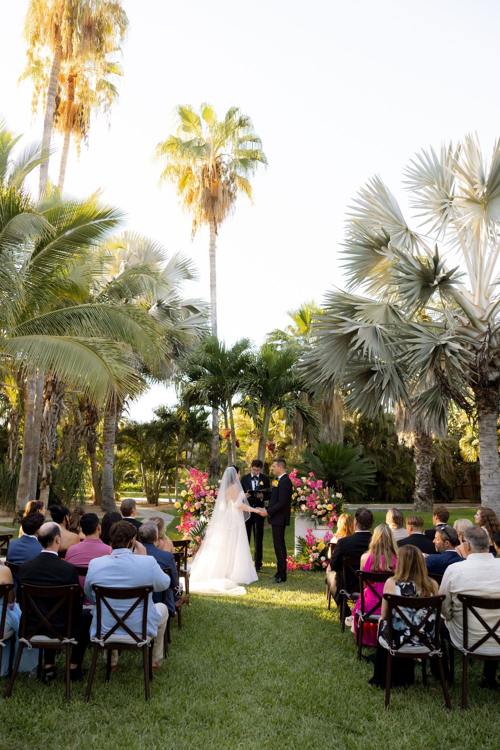 Outdoor tropical wedding ceremony with bride and groom at floral altar surrounded by palm trees.
