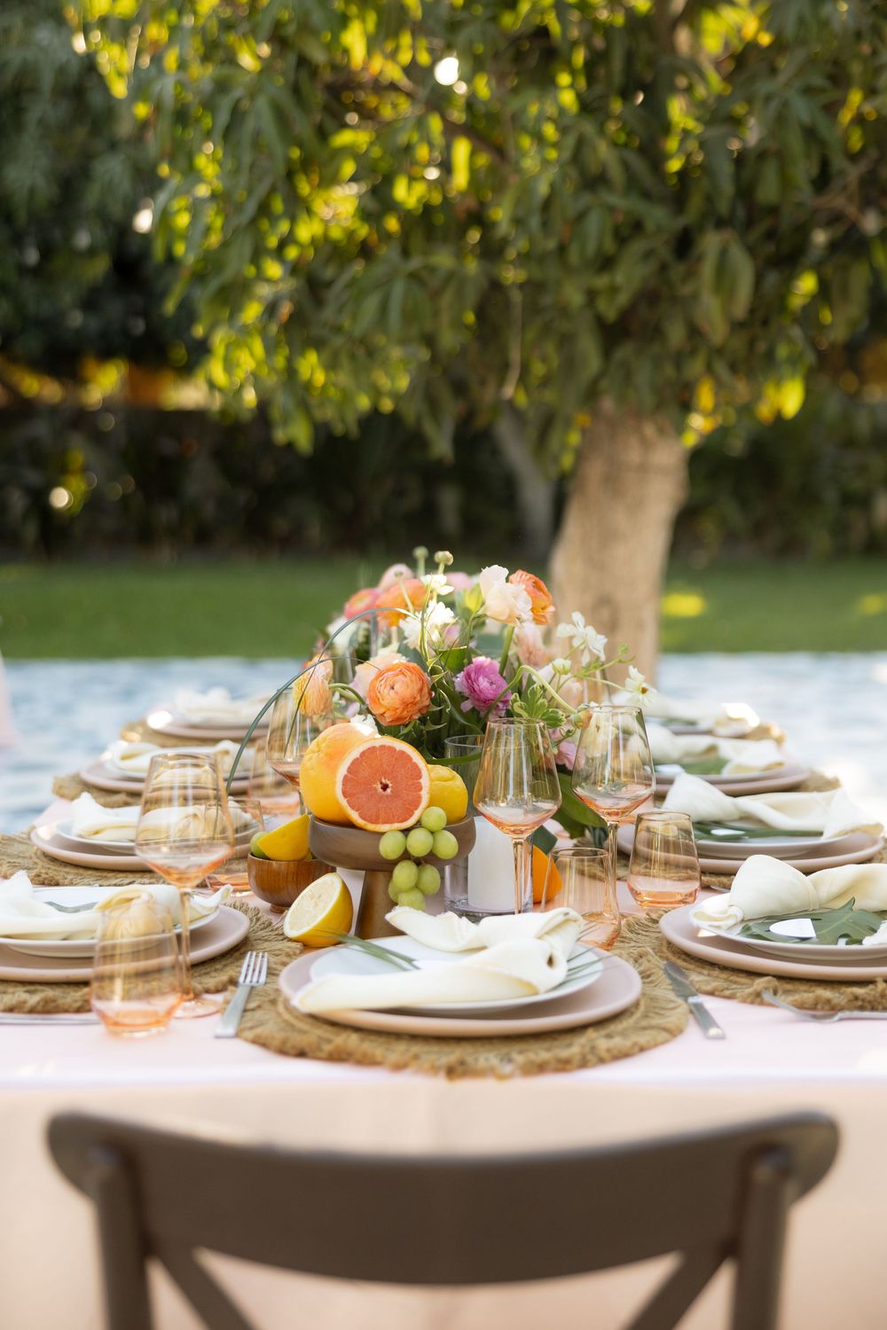 Outdoor wedding reception table set with citrus fruit centerpiece, wine glasses, and linen napkins under a green tree.