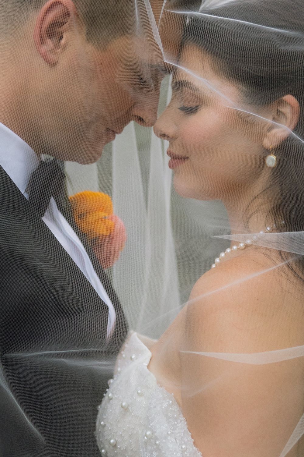 Intimate close-up of bride and groom touching foreheads tenderly under a sheer wedding veil with orange boutonniere.