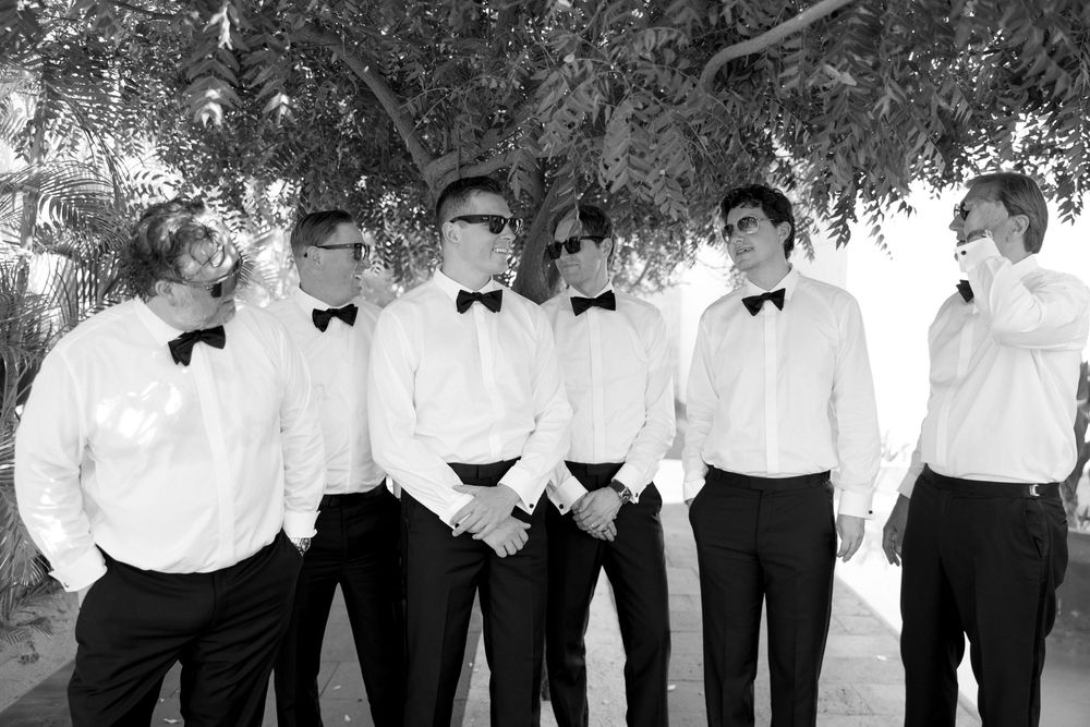 Black and white photo of groomsmen in tuxedos and sunglasses gathered under a shady tree.