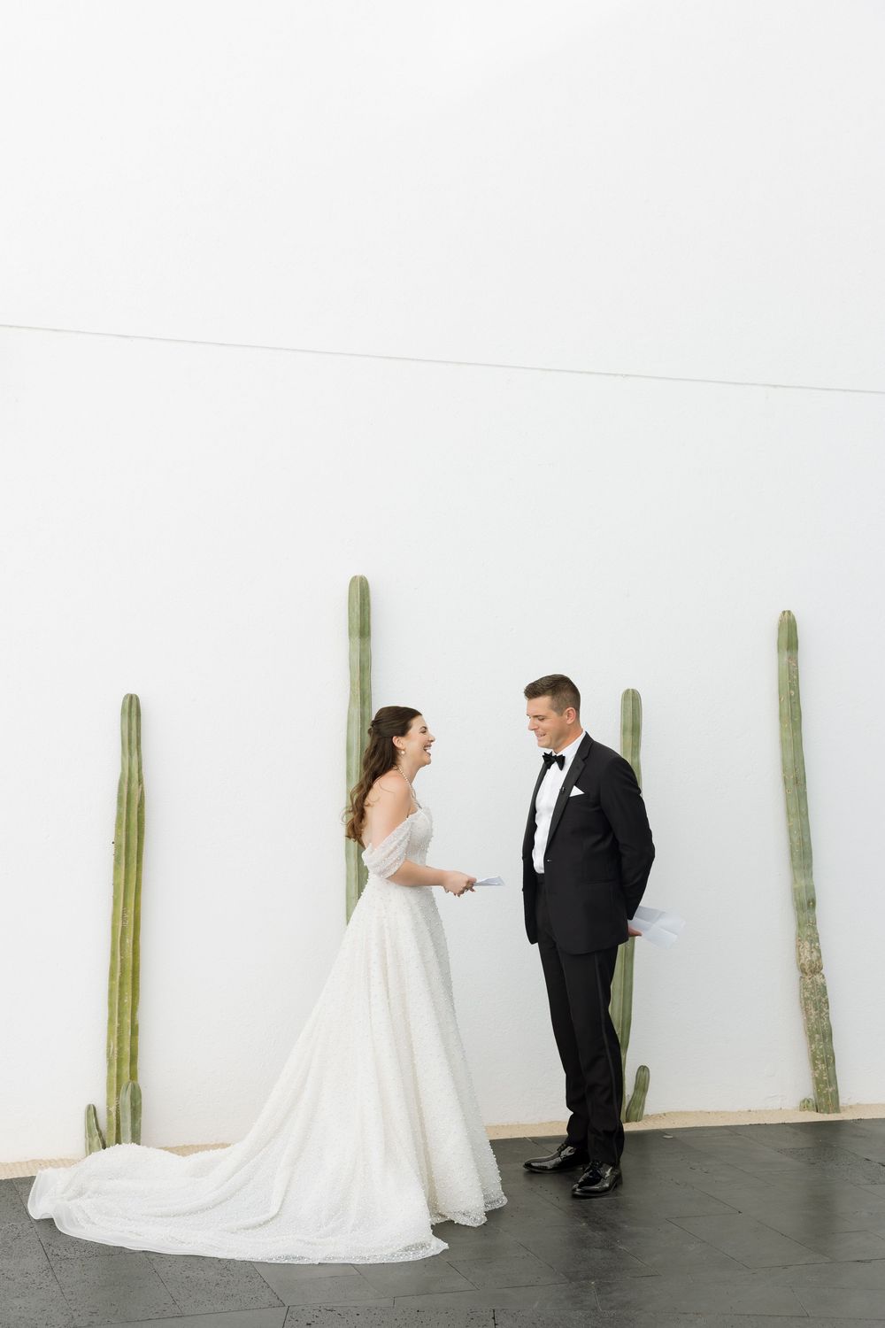 Bride and groom exchange vows in front of white wall with tall cacti at modern wedding venue.