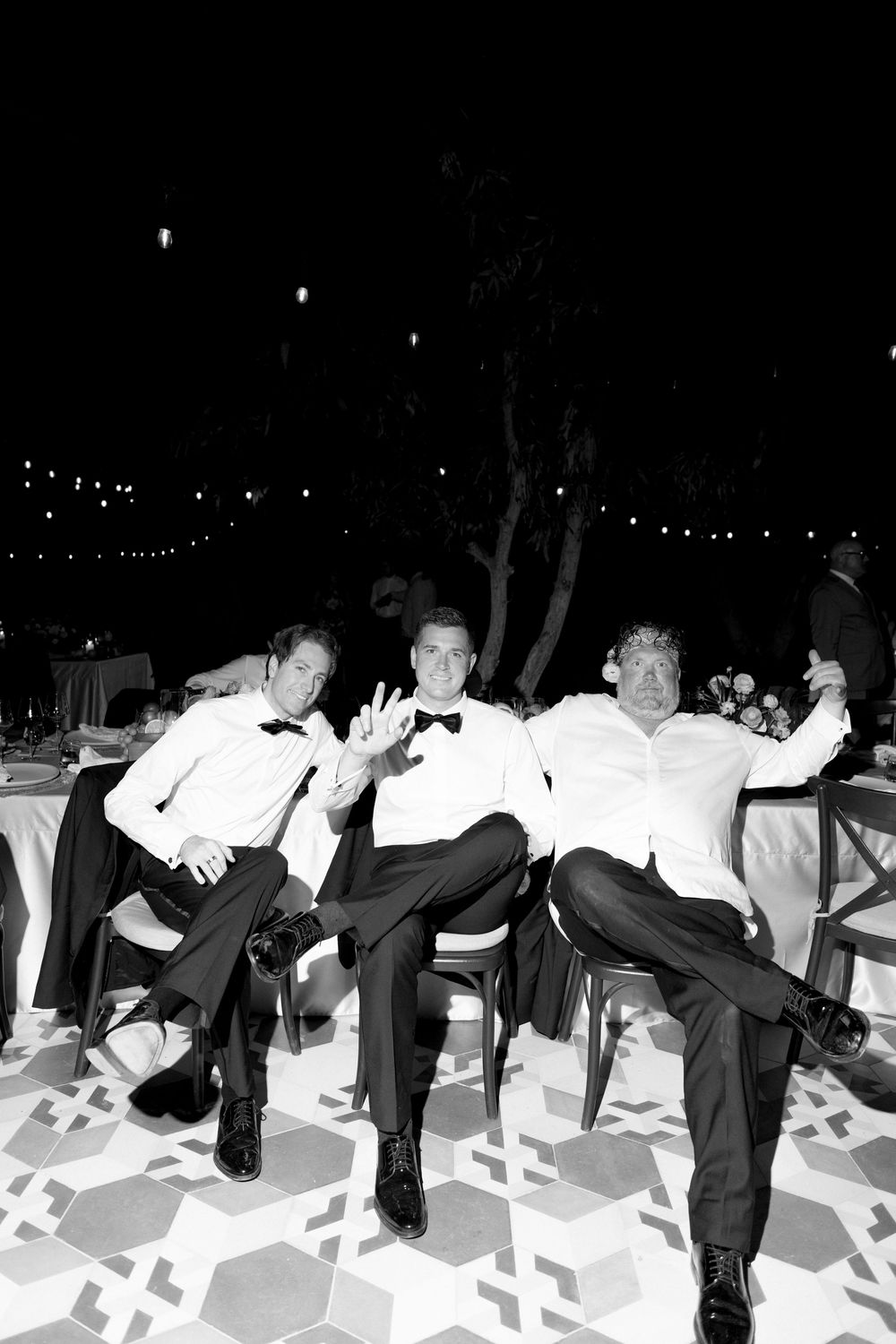 Three groomsmen in tuxedos pose playfully seated at a wedding reception table in a black and white photo.