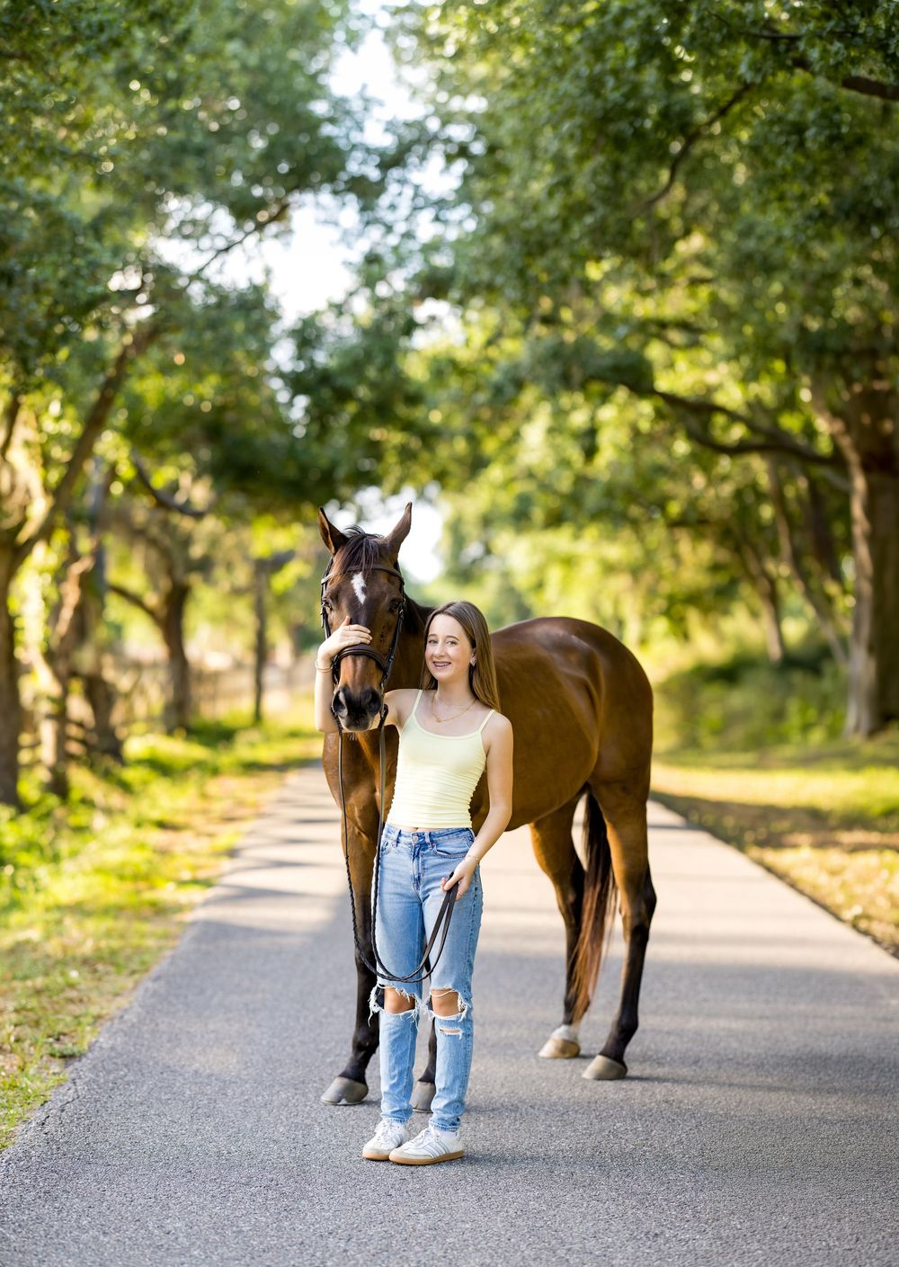 Young woman in jeans and yellow top walks a chestnut horse down a tree-lined path on a sunny day.