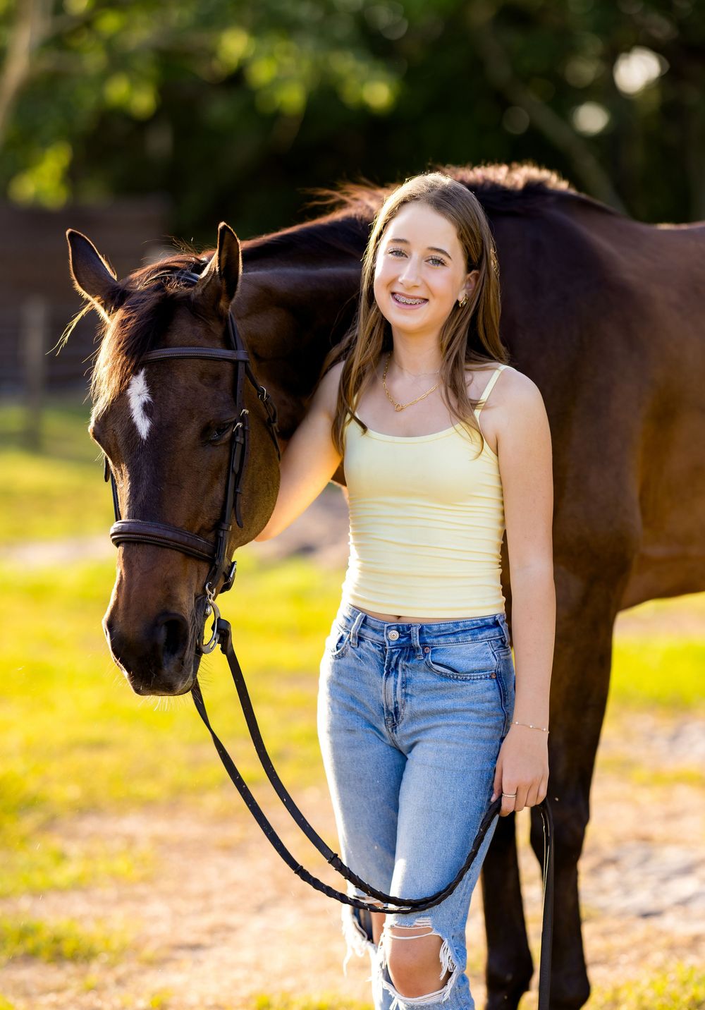 Young woman in yellow top and jeans smiling beside a dark brown horse on a sunny green field.