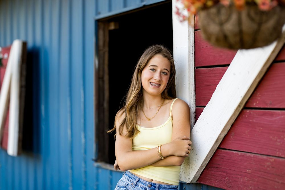 Smiling teenage girl with braces in yellow top leaning against a rustic red and blue barn door.