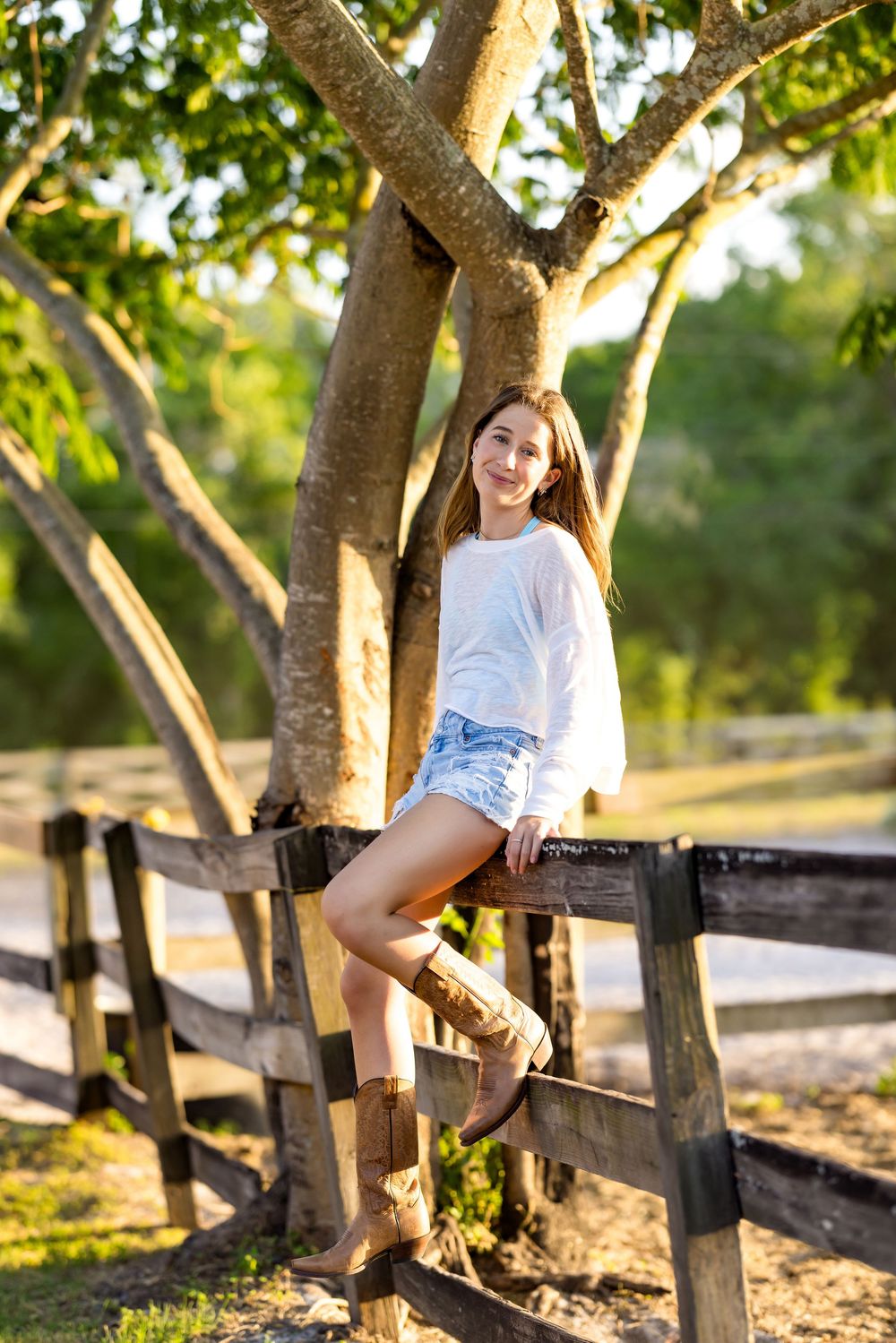 Young woman in white top, denim shorts, and cowboy boots sitting on a rustic wooden fence outdoors.