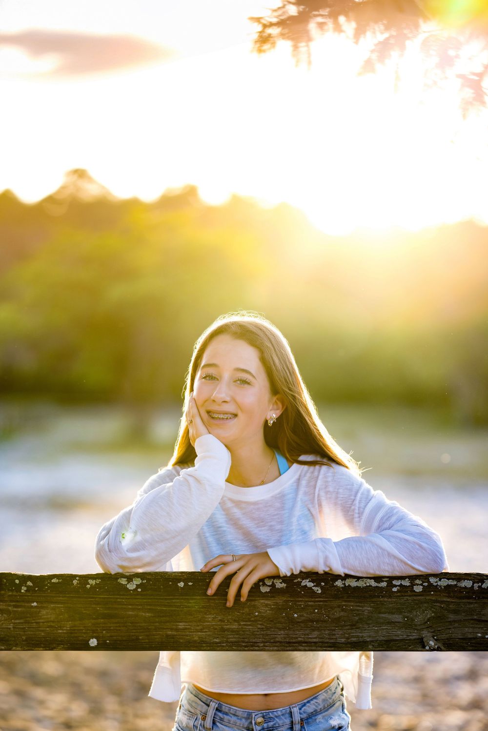 Young woman leaning on wooden fence at golden hour sunset, smiling outdoors in white crop top and jeans.