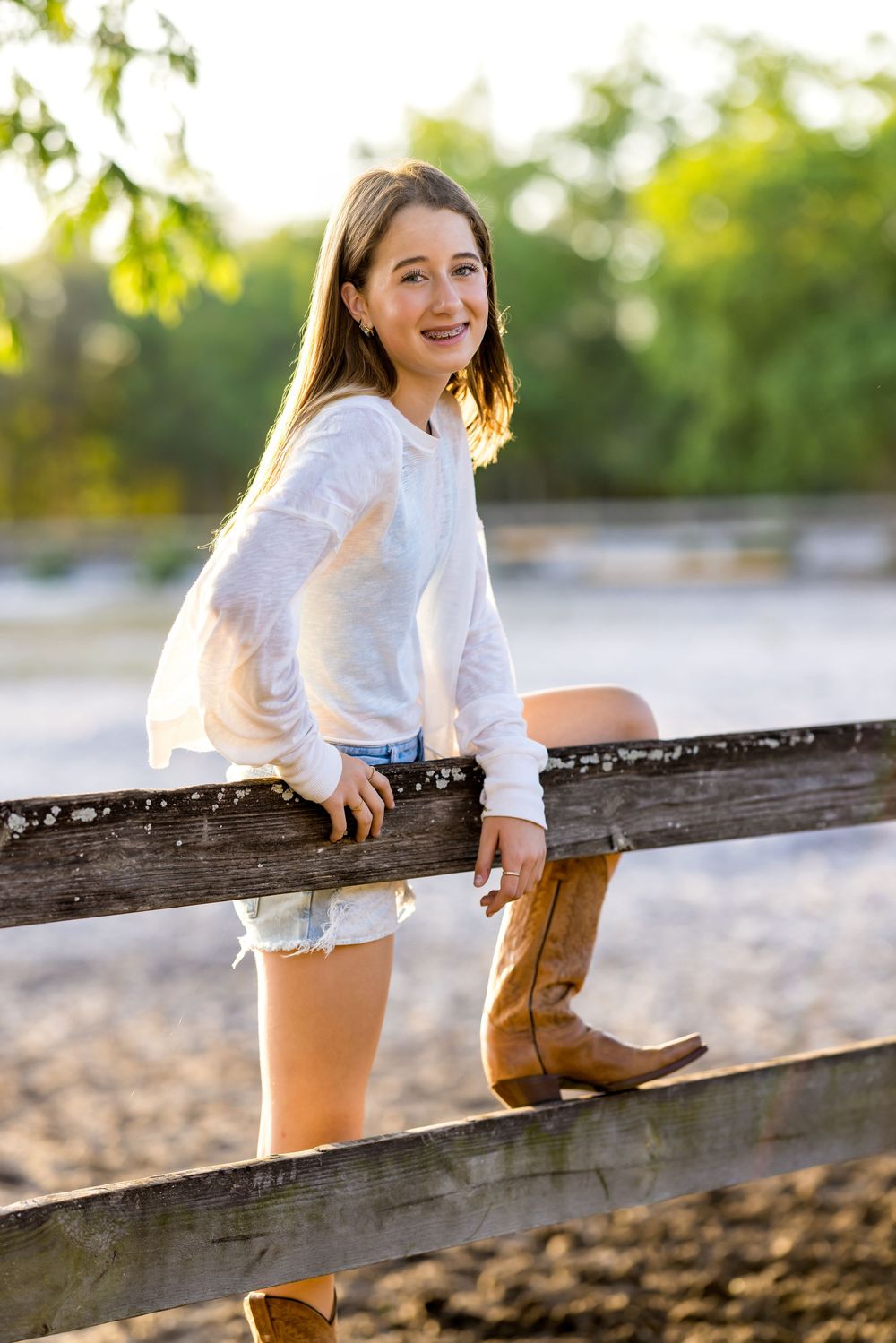 Young girl in white top, denim shorts, and cowboy boots leaning on a wooden fence outdoors.