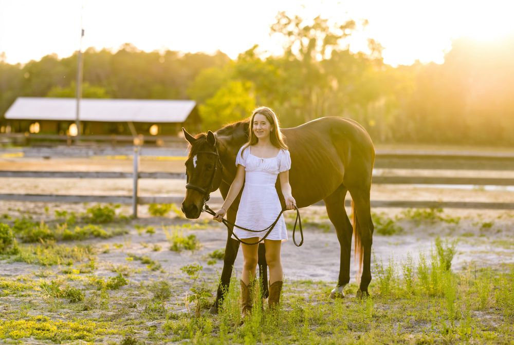 Young woman in white dress and cowboy boots leading a dark horse at golden hour on a ranch.