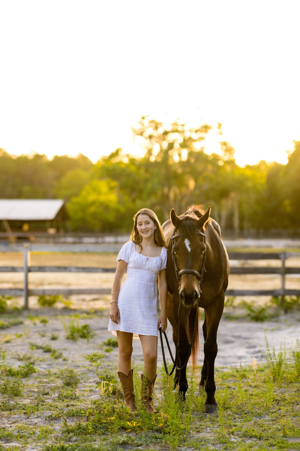 A smiling girl in a white dress and cowboy boots stands beside a dark brown horse in a sunny farm setting.