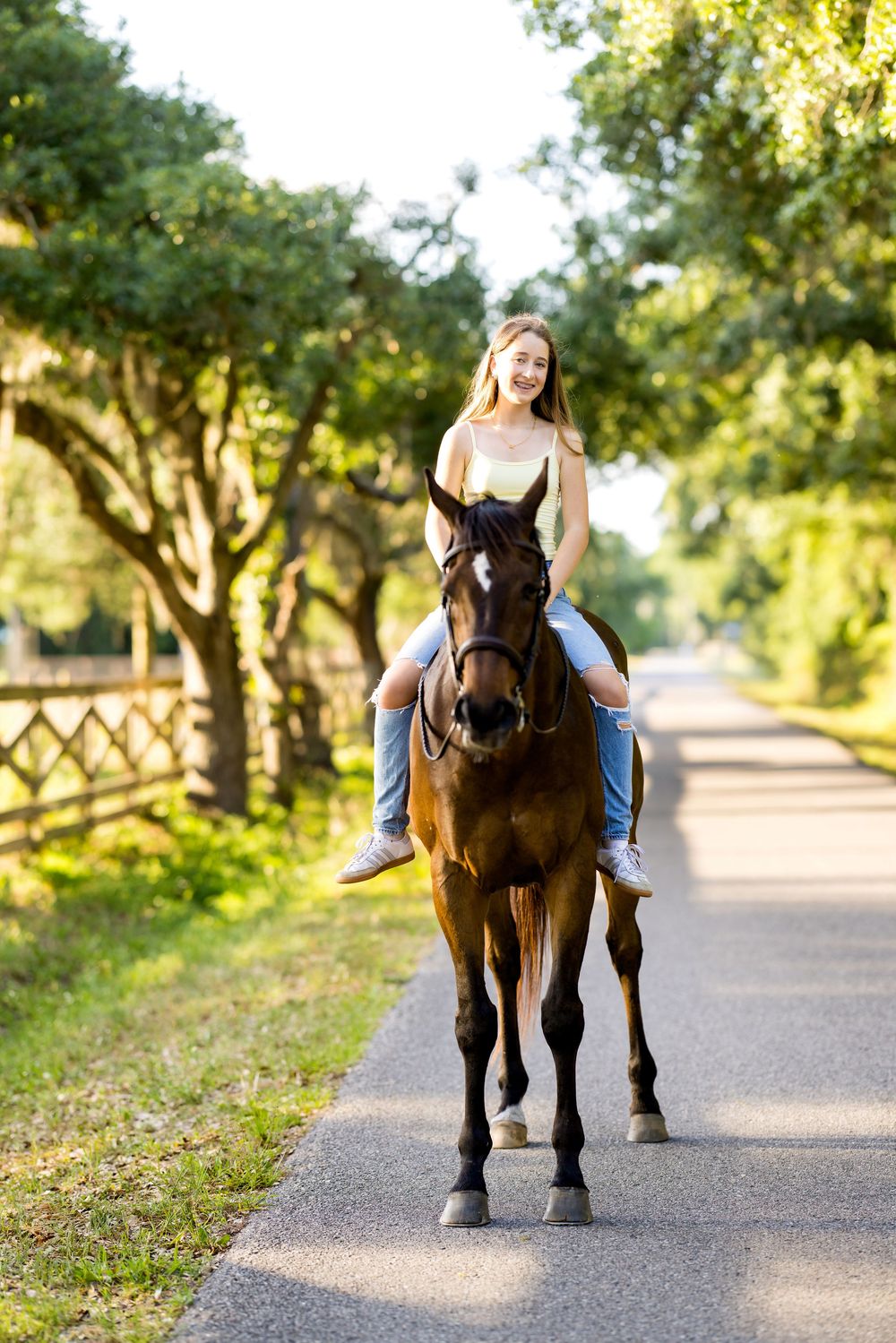 Young woman riding a brown horse on a sunny tree-lined path in a lush green park setting.