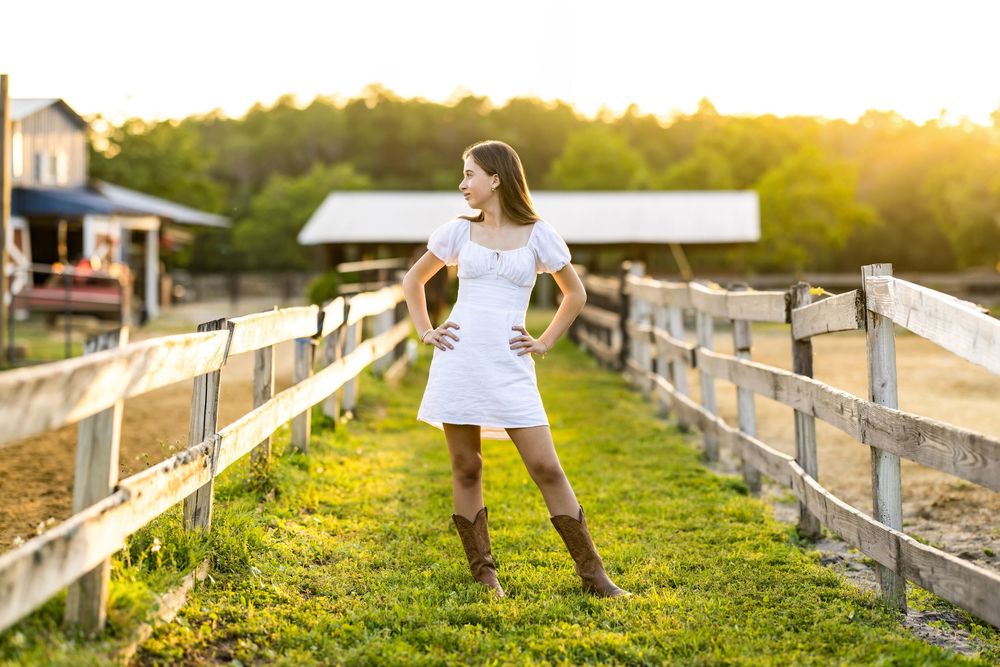 Young woman in white dress and brown boots poses by wooden fence on a sunny farm at golden hour.