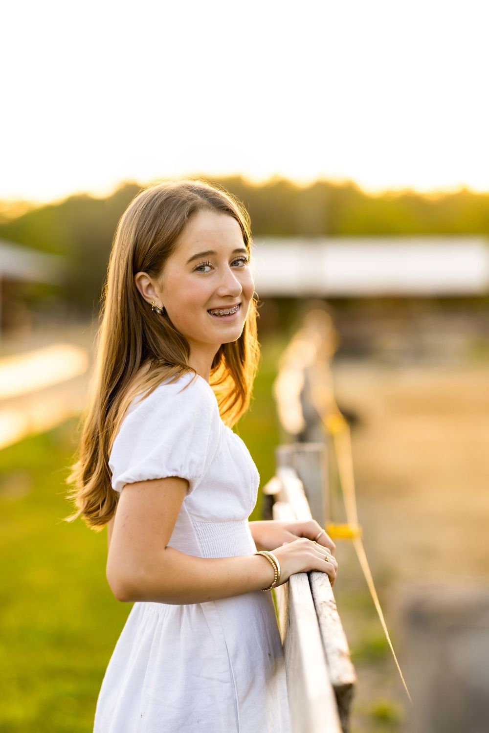 Young girl in white dress smiling at camera while leaning on wooden fence at golden hour sunset.