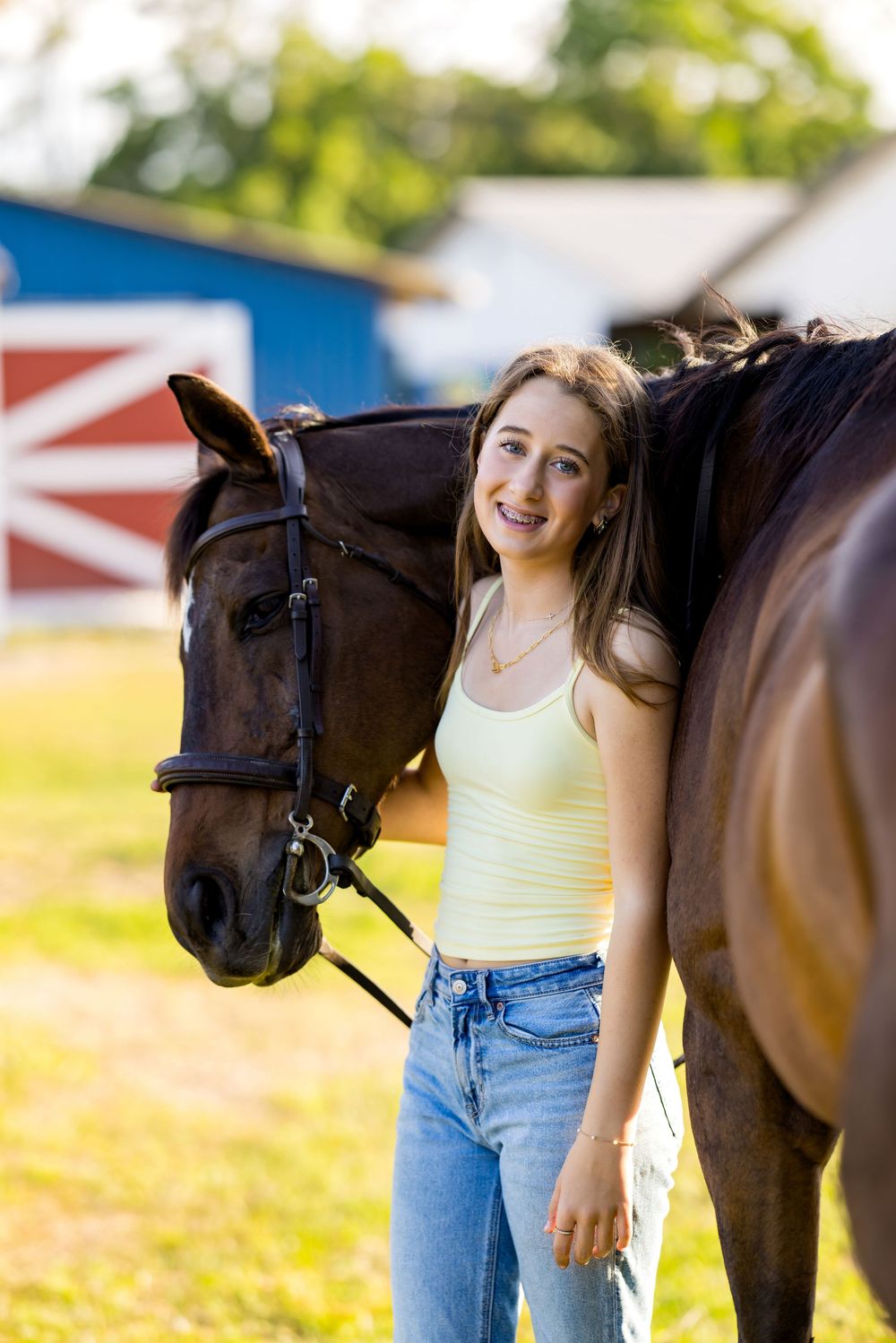Young woman in yellow tank top and jeans smiling beside a brown horse near a red barn.