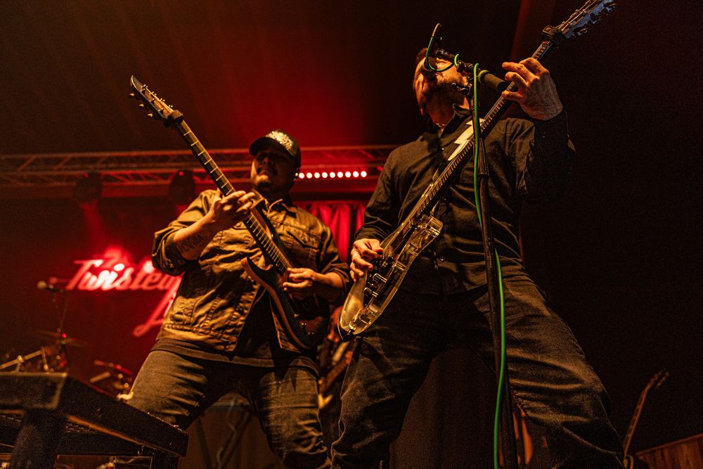 Two guitarists rock out on a darkly lit stage with red and green lighting.