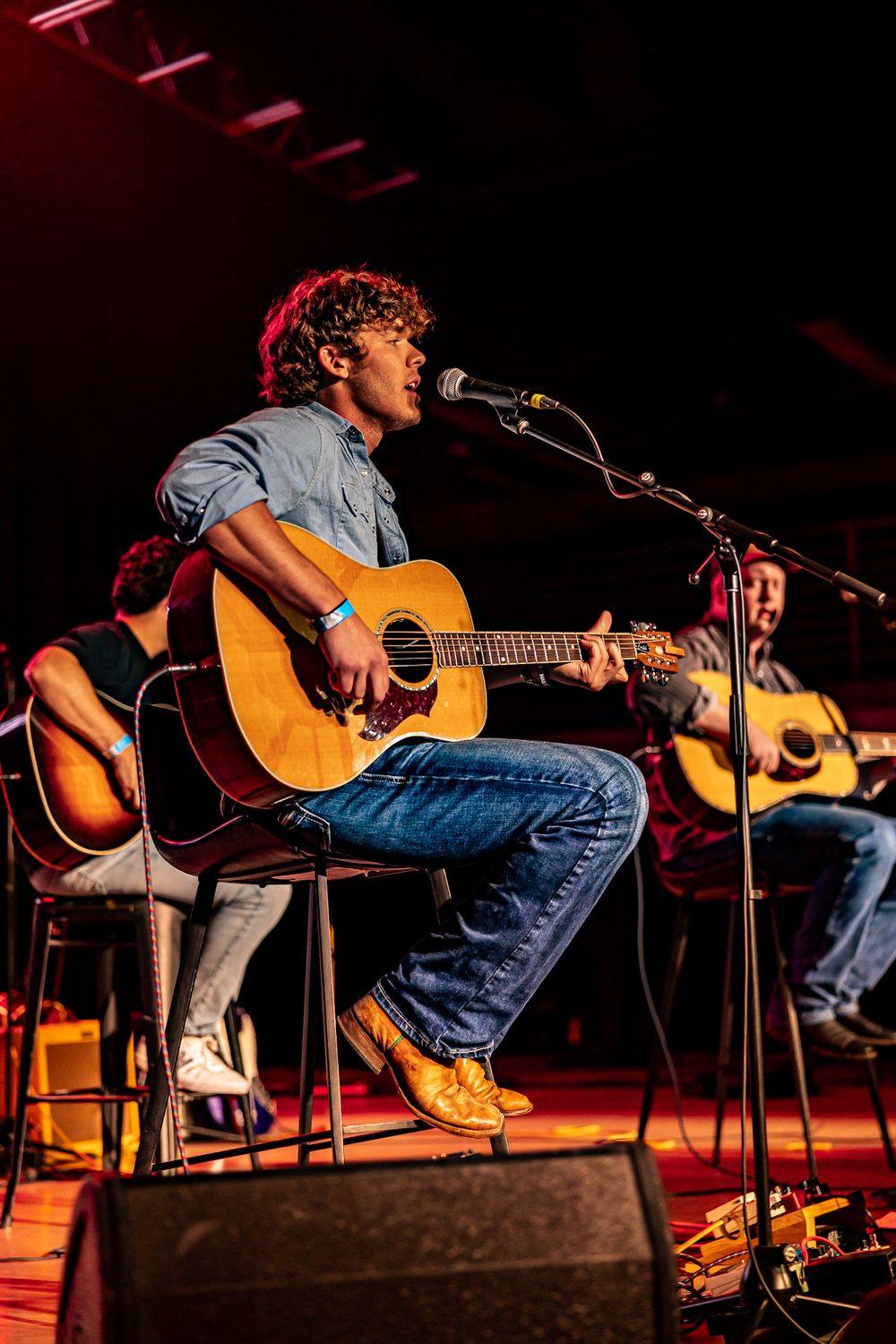 Young male guitarist sings into microphone during acoustic performance on stage.