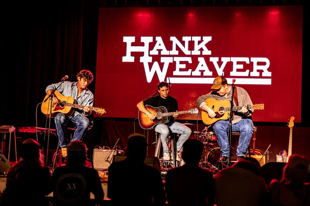 Three acoustic guitarists perform under Hank Weaver sign on a red-lit stage.