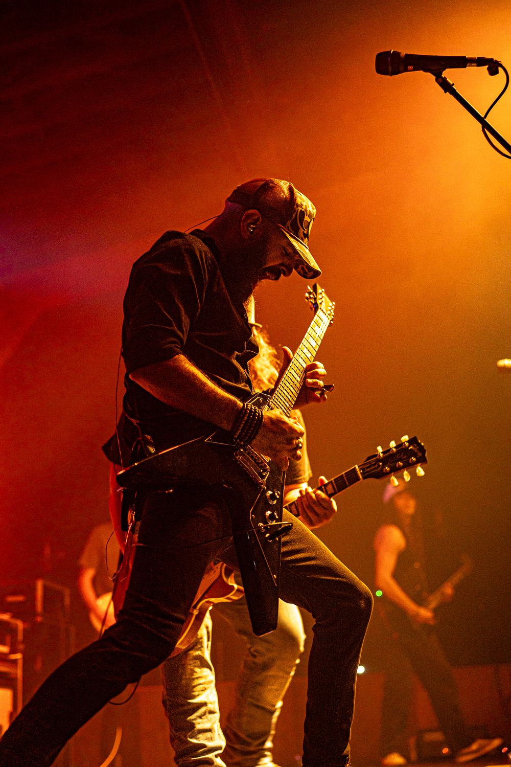 Male guitarist plays electric guitar on stage under dramatic orange and red concert lighting.