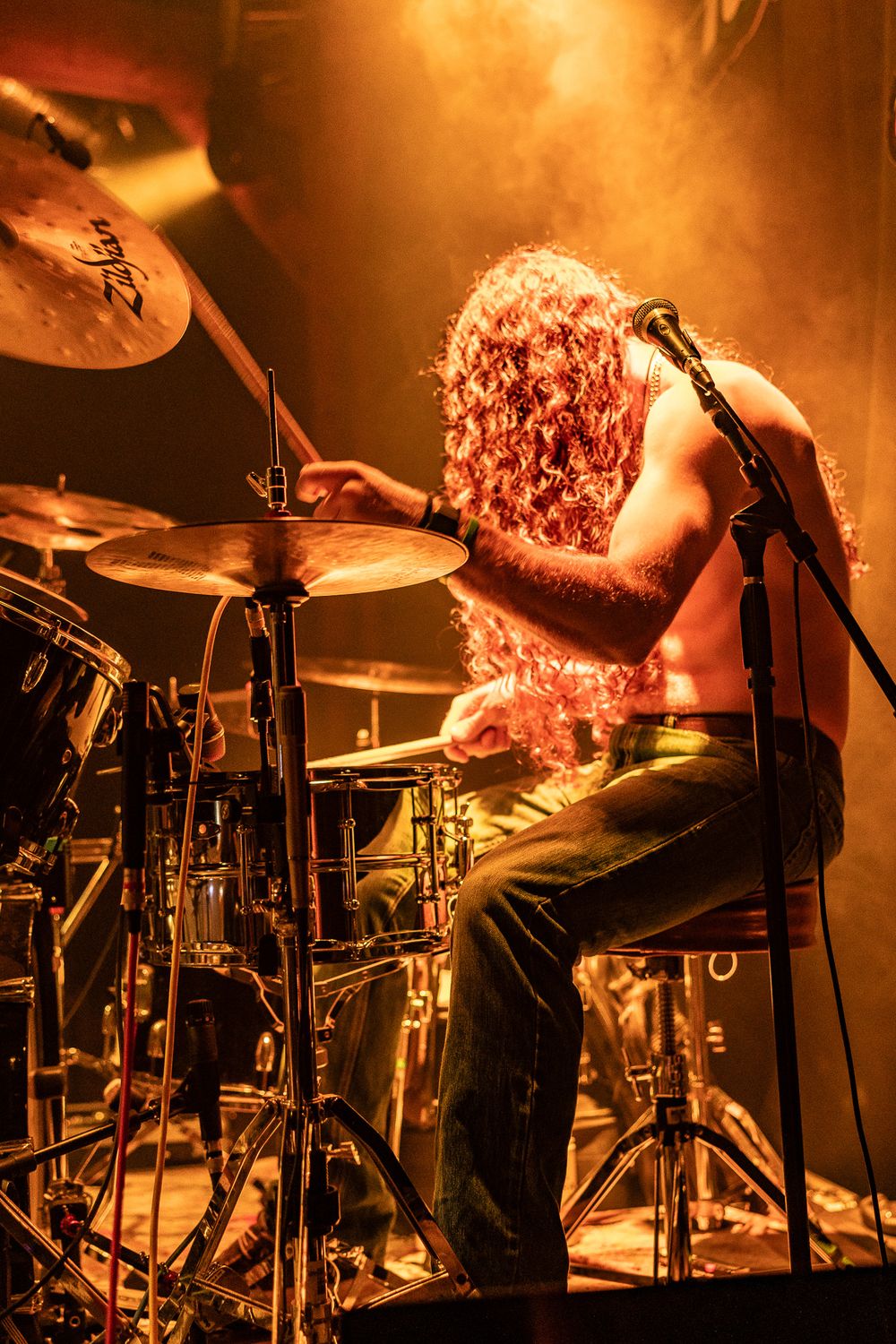 Shirtless drummer with long curly hair plays Zildjian cymbals on stage under warm orange lights.