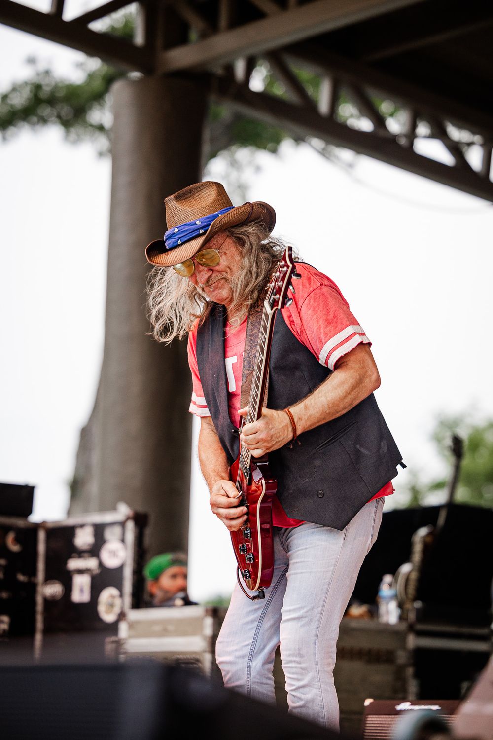 Long-haired musician in cowboy hat plays electric guitar on outdoor stage during daytime concert.