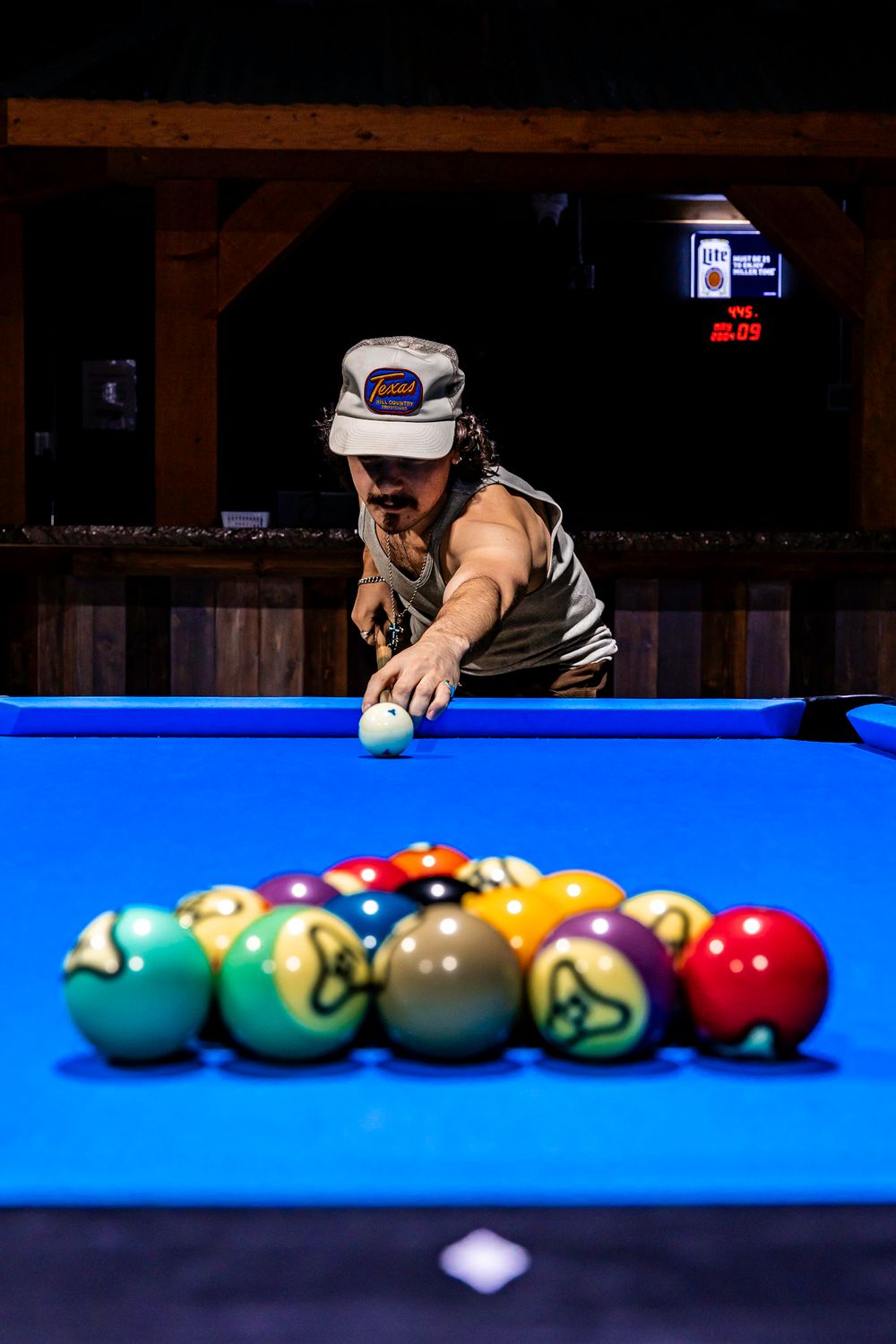 Man in trucker hat shoots pool on a blue billiard table in a rustic bar.