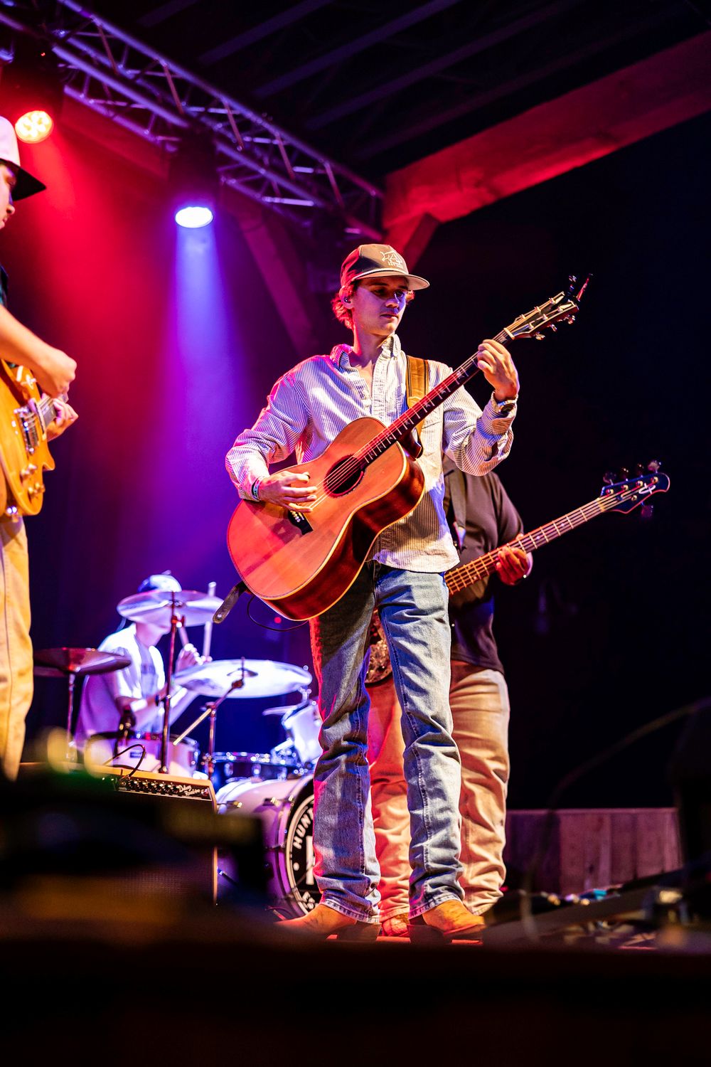 Young guitarist in cap plays acoustic guitar on a colorful lit stage during a live concert performance.